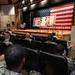 A military briefing or ceremony is taking place in an auditorium, with a large audience of uniformed Navy personnel seated in rows facing a stage. The stage is decorated with a large American flag as a backdrop, along with display boards containing photographs and text. A speaker stands at a podium on stage while a Navy petty officer in dress whites stands at attention near a ship's bell on the right side.