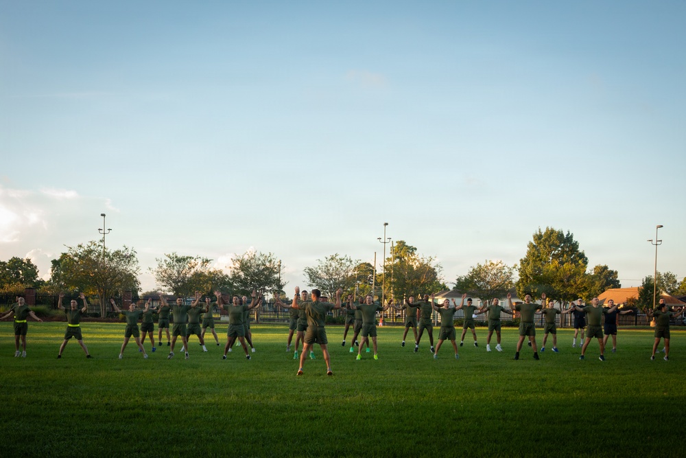 A large group of individuals dressed in matching olive/dark green military-style physical training uniforms are performing synchronized exercises on a well-maintained grass field. They appear to be doing jumping jacks or a similar calisthenic movement, led by a central figure facing the group. The scene is set during golden hour lighting with trees, light poles, and a fenced perimeter visible in the background.