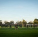 A large group of individuals dressed in matching olive/dark green military-style physical training uniforms are performing synchronized exercises on a well-maintained grass field. They appear to be doing jumping jacks or a similar calisthenic movement, led by a central figure facing the group. The scene is set during golden hour lighting with trees, light poles, and a fenced perimeter visible in the background.