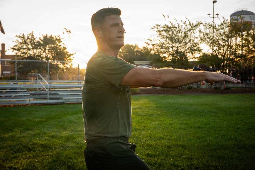 A man in an olive green t-shirt and dark pants is performing a stretching or exercise movement outdoors on a grass field, with his arms extended forward. The scene is backlit by a golden sunset, creating warm lens flare effects. The setting appears to be a sports or recreational facility with bleachers, fencing, trees, and a water tower visible in the background.