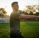 A man in an olive green t-shirt and dark pants is performing a stretching or exercise movement outdoors on a grass field, with his arms extended forward. The scene is backlit by a golden sunset, creating warm lens flare effects. The setting appears to be a sports or recreational facility with bleachers, fencing, trees, and a water tower visible in the background.