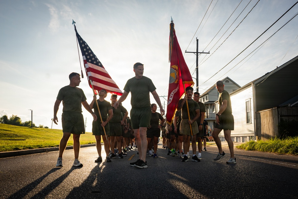 A group of U.S. Marines in physical training (PT) uniforms — olive green t-shirts and shorts — march or run in formation along a residential street. Two members carry flags: the United States flag and the U.S. Marine Corps flag. The scene appears to take place during early morning or late afternoon based on the low-angle golden sunlight.