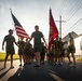 A group of U.S. Marines in physical training (PT) uniforms — olive green t-shirts and shorts — march or run in formation along a residential street. Two members carry flags: the United States flag and the U.S. Marine Corps flag. The scene appears to take place during early morning or late afternoon based on the low-angle golden sunlight.