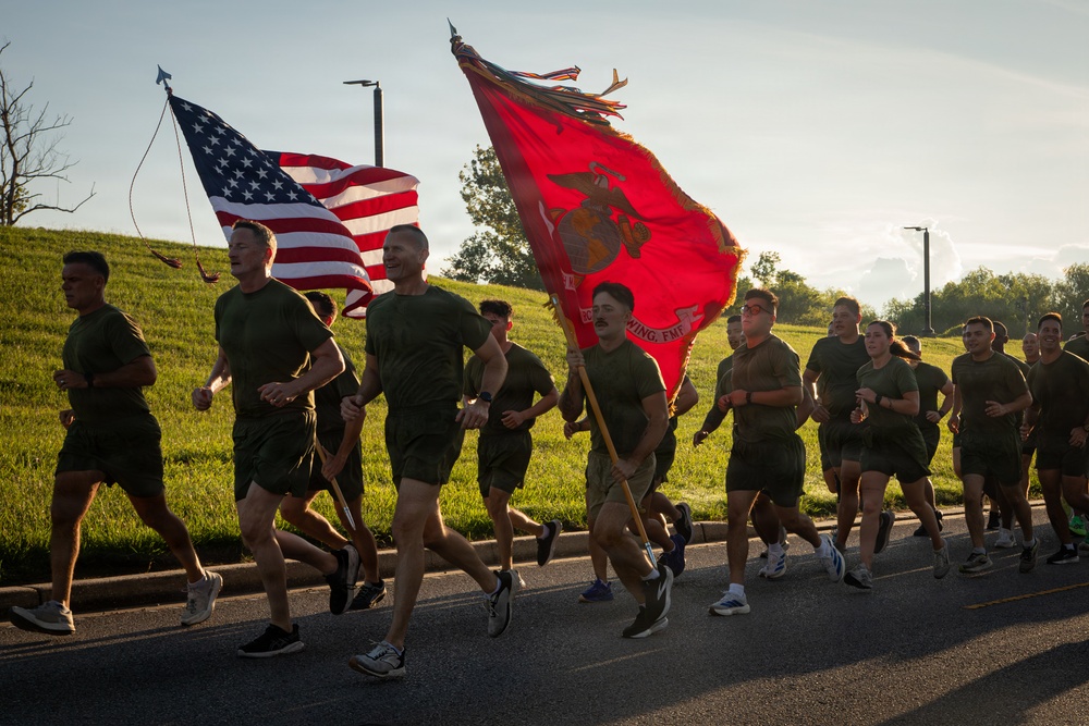 A large group of U.S. Marines in physical training (PT) uniforms — olive green t-shirts and shorts — are running in formation along a paved road. Two flag bearers carry the American flag and a red U.S. Marine Corps unit guidon with the Eagle, Globe, and Anchor emblem. The scene appears to take place during early morning hours based on the low-angle sunlight and warm golden lighting.