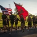 A large group of U.S. Marines in physical training (PT) uniforms — olive green t-shirts and shorts — are running in formation along a paved road. Two flag bearers carry the American flag and a red U.S. Marine Corps unit guidon with the Eagle, Globe, and Anchor emblem. The scene appears to take place during early morning hours based on the low-angle sunlight and warm golden lighting.