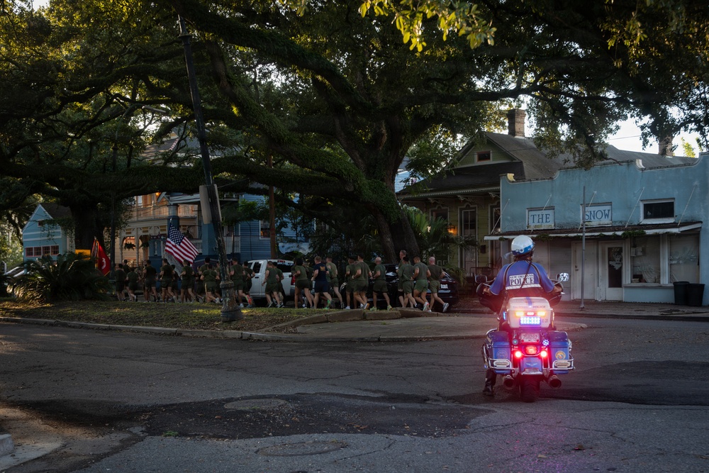 A group of uniformed runners, appearing to be military personnel, jog in formation along a tree-lined residential street while carrying American flags and what appears to be a military unit flag. A police officer on a motorcycle escorts the group, with emergency lights activated. The scene appears to take place in a Southern U.S. city, likely New Orleans, based on the architectural style of the buildings.