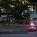 A group of uniformed runners, appearing to be military personnel, jog in formation along a tree-lined residential street while carrying American flags and what appears to be a military unit flag. A police officer on a motorcycle escorts the group, with emergency lights activated. The scene appears to take place in a Southern U.S. city, likely New Orleans, based on the architectural style of the buildings.