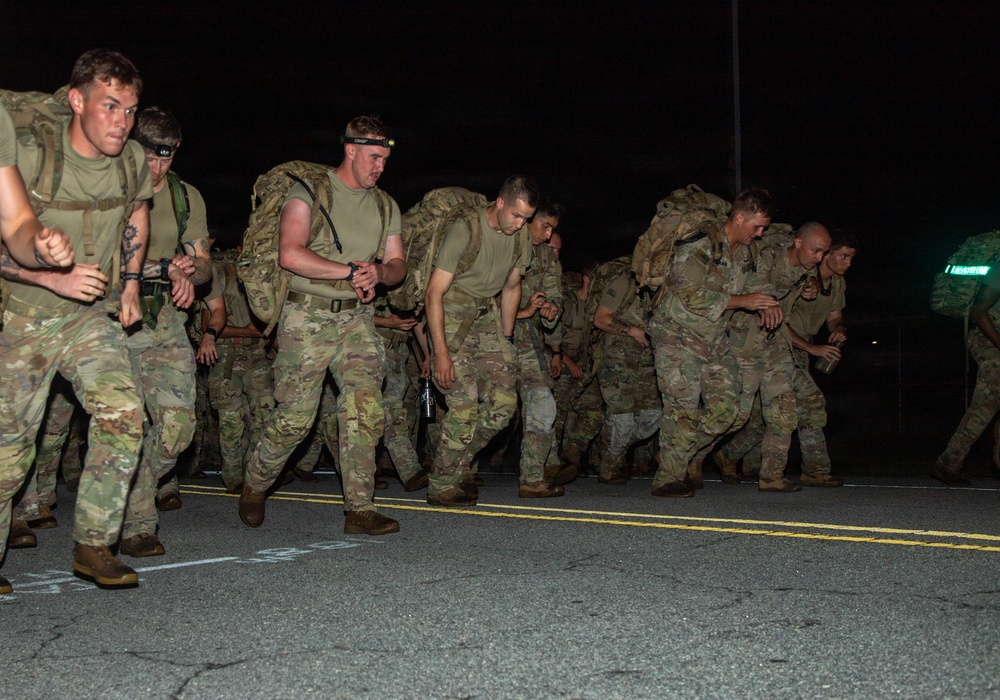 A group of military personnel in camouflage uniforms are conducting a ruck march or physical training exercise at night on a paved road. The soldiers are carrying heavy rucksacks on their backs and appear to be moving at a fast pace, leaning forward under the weight of their gear. The scene is lit primarily by ambient light, with some soldiers wearing headlamps.