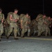 A group of military personnel in camouflage uniforms are conducting a ruck march or physical training exercise at night on a paved road. The soldiers are carrying heavy rucksacks on their backs and appear to be moving at a fast pace, leaning forward under the weight of their gear. The scene is lit primarily by ambient light, with some soldiers wearing headlamps.