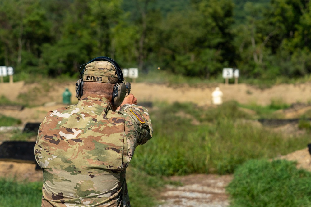 A military service member named Watkins is firing a pistol at a shooting range. The soldier is wearing OCP (Operational Camouflage Pattern) uniform with hearing protection and is engaged in marksmanship training. Multiple target stands are visible downrange in an outdoor range setting.