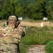 A military service member named Watkins is firing a pistol at a shooting range. The soldier is wearing OCP (Operational Camouflage Pattern) uniform with hearing protection and is engaged in marksmanship training. Multiple target stands are visible downrange in an outdoor range setting.