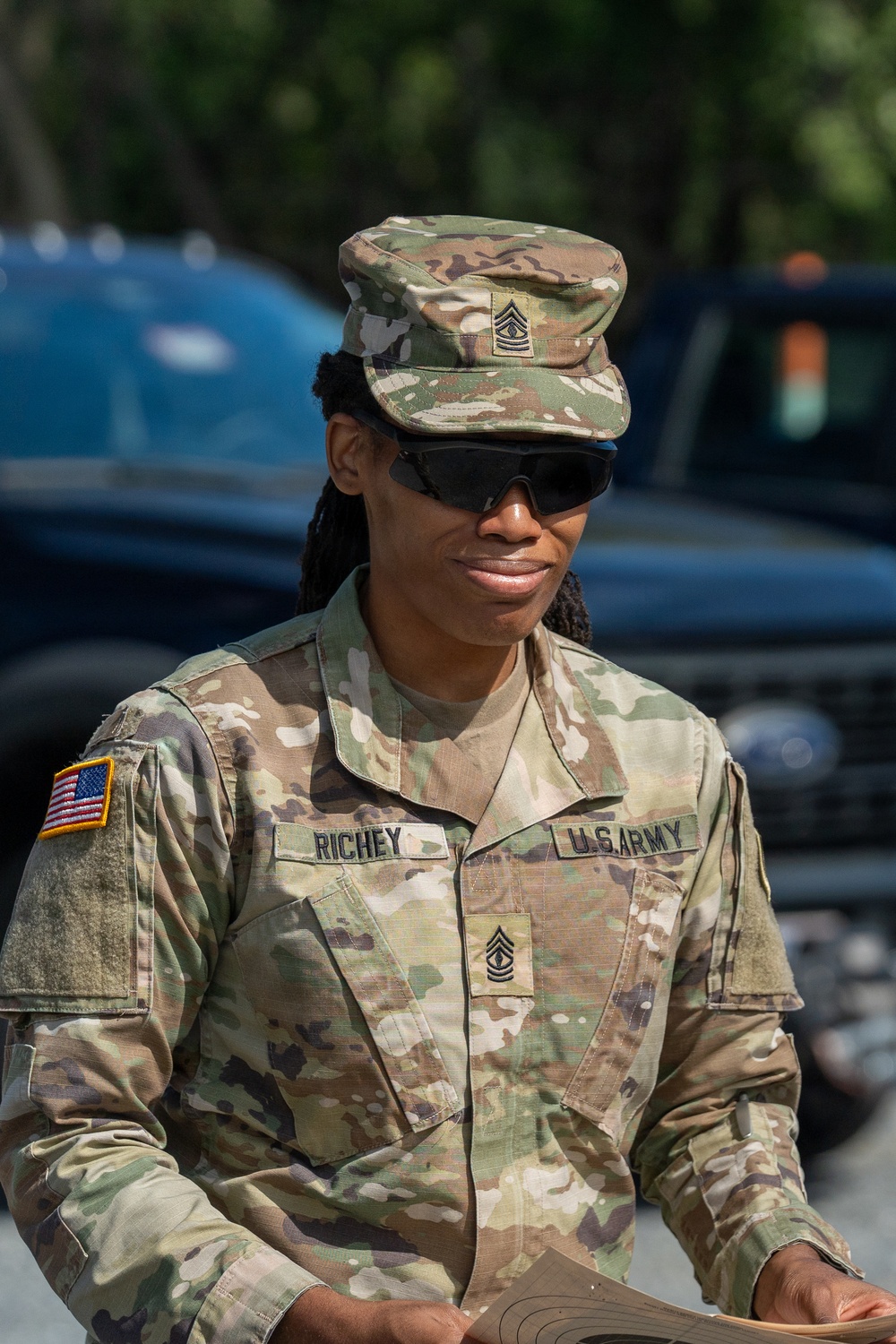 A U.S. Army soldier wearing Operational Camouflage Pattern (OCP) uniform is photographed outdoors. The soldier, identified by name tape as 'RICHEY,' holds the rank of Sergeant Major as indicated by insignia on the patrol cap and chest. The soldier is wearing ballistic/protective eyewear and appears to be reviewing a document.