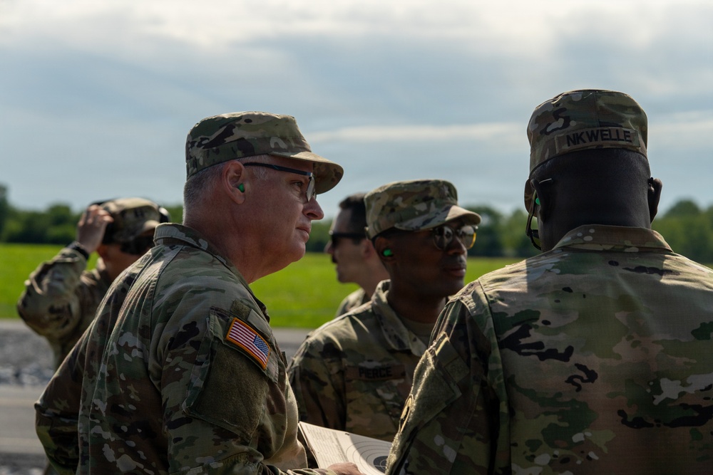 A group of U.S. Army soldiers in OCP (Operational Camouflage Pattern) uniforms are gathered outdoors, appearing to be in a briefing or review session. A senior soldier on the left is holding what appears to be a document or target sheet while engaging with fellow soldiers. The setting appears to be a training range or airfield based on the open terrain and paved surface visible.