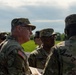 A group of U.S. Army soldiers in OCP (Operational Camouflage Pattern) uniforms are gathered outdoors, appearing to be in a briefing or review session. A senior soldier on the left is holding what appears to be a document or target sheet while engaging with fellow soldiers. The setting appears to be a training range or airfield based on the open terrain and paved surface visible.