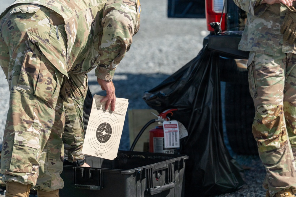Two military personnel in OCP (Operational Camouflage Pattern) uniforms are handling equipment near what appears to be a vehicle. One soldier is holding a circular shooting target paper, while a black plastic storage case and a fire extinguisher are visible nearby. The scene appears to be associated with a marksmanship or weapons qualification activity.