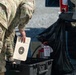 Two military personnel in OCP (Operational Camouflage Pattern) uniforms are handling equipment near what appears to be a vehicle. One soldier is holding a circular shooting target paper, while a black plastic storage case and a fire extinguisher are visible nearby. The scene appears to be associated with a marksmanship or weapons qualification activity.