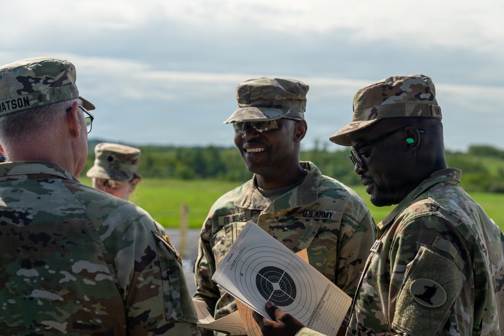 Several U.S. Army soldiers in OCP (Operational Camouflage Pattern) uniforms are gathered outdoors, engaged in conversation at what appears to be a shooting range. One soldier is holding a paper shooting target with a bullseye pattern. The setting appears to be a training exercise or qualification event in a rural, open field environment.