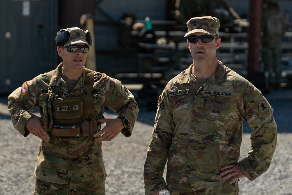 Two U.S. Army soldiers in Operational Camouflage Pattern (OCP) uniforms are standing outdoors at what appears to be a military installation or training area. The soldier on the left, identified by name tape as 'WESTERVELT,' is wearing a tactical chest rig and hearing protection pushed up on his head. The soldier on the right, identified as 'SULLIVAN,' wears a major's rank insignia (gold oak leaf) and a '1st Infantry Division' patch.