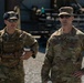 Two U.S. Army soldiers in Operational Camouflage Pattern (OCP) uniforms are standing outdoors at what appears to be a military installation or training area. The soldier on the left, identified by name tape as 'WESTERVELT,' is wearing a tactical chest rig and hearing protection pushed up on his head. The soldier on the right, identified as 'SULLIVAN,' wears a major's rank insignia (gold oak leaf) and a '1st Infantry Division' patch.