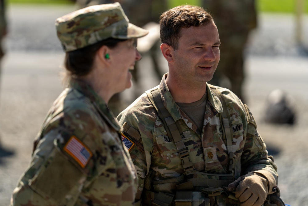 Two U.S. Army soldiers in Operational Camouflage Pattern (OCP) uniforms are engaged in conversation. Both wear American flag patches on their left shoulders, indicating active U.S. military personnel. The male soldier on the right has a partially visible name tape and a rank insignia (appears to be Major) on his chest.