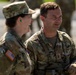 Two U.S. Army soldiers in Operational Camouflage Pattern (OCP) uniforms are engaged in conversation. Both wear American flag patches on their left shoulders, indicating active U.S. military personnel. The male soldier on the right has a partially visible name tape and a rank insignia (appears to be Major) on his chest.