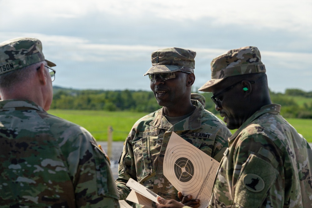 Three U.S. Army soldiers in OCP (Operational Camouflage Pattern) uniforms are engaged in conversation at what appears to be a shooting range. The central soldier is holding a paper shooting target with concentric circles, suggesting a marksmanship training or qualification event. The setting is an outdoor range with green fields and trees visible in the background.