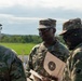 Three U.S. Army soldiers in OCP (Operational Camouflage Pattern) uniforms are engaged in conversation at what appears to be a shooting range. The central soldier is holding a paper shooting target with concentric circles, suggesting a marksmanship training or qualification event. The setting is an outdoor range with green fields and trees visible in the background.