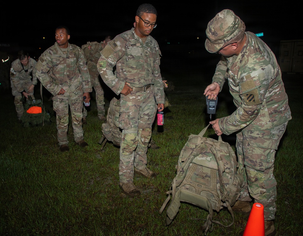 A group of U.S. Army soldiers in OCP (Operational Camouflage Pattern) uniforms are lined up at night on a grass field. A soldier on the right is using a handheld digital scale to weigh a tactical backpack, likely conducting a ruck weight inspection before a march or training event. Other soldiers are waiting in line, some carrying gear and personal items.