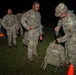 A group of U.S. Army soldiers in OCP (Operational Camouflage Pattern) uniforms are lined up at night on a grass field. A soldier on the right is using a handheld digital scale to weigh a tactical backpack, likely conducting a ruck weight inspection before a march or training event. Other soldiers are waiting in line, some carrying gear and personal items.