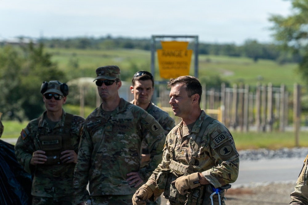 A group of U.S. Army soldiers in MultiCam/OCP camouflage uniforms are gathered outdoors near a military range area. The foreground soldier displays an EOD (Explosive Ordnance Disposal) tab patch on his uniform. The setting appears to be a training range facility, indicated by a yellow 'Range 4' sign visible in the background.