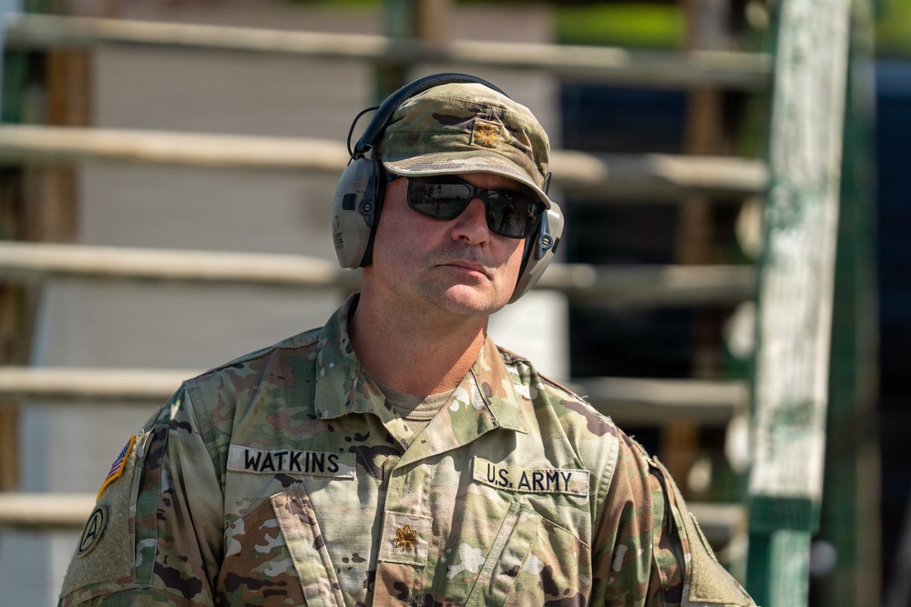 A U.S. Army soldier identified by name tape as 'Watkins' is photographed in a close-up portrait-style image. He is wearing OCP (Operational Camouflage Pattern) uniform with a major's oak leaf insignia, tactical hearing protection headset, sunglasses, and a patrol cap. The setting appears to be an outdoor range or training environment.