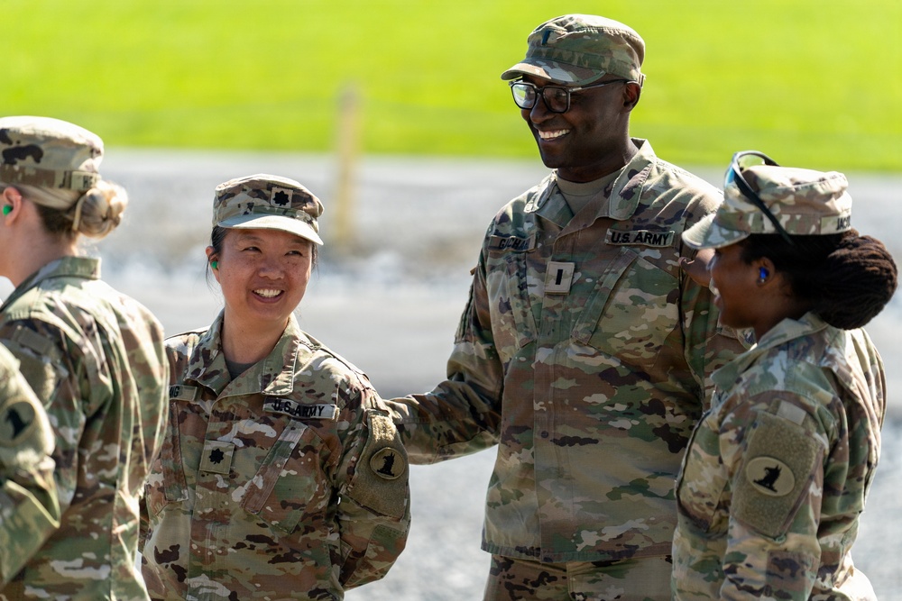 A group of U.S. Army soldiers in OCP (Operational Camouflage Pattern) uniforms are engaged in a friendly interaction outdoors. At least four soldiers are visible, with two in the center smiling and appearing to share a positive exchange. The setting appears to be a military installation or training area with a paved surface and green grass in the background.