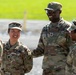 A group of U.S. Army soldiers in OCP (Operational Camouflage Pattern) uniforms are engaged in a friendly interaction outdoors. At least four soldiers are visible, with two in the center smiling and appearing to share a positive exchange. The setting appears to be a military installation or training area with a paved surface and green grass in the background.