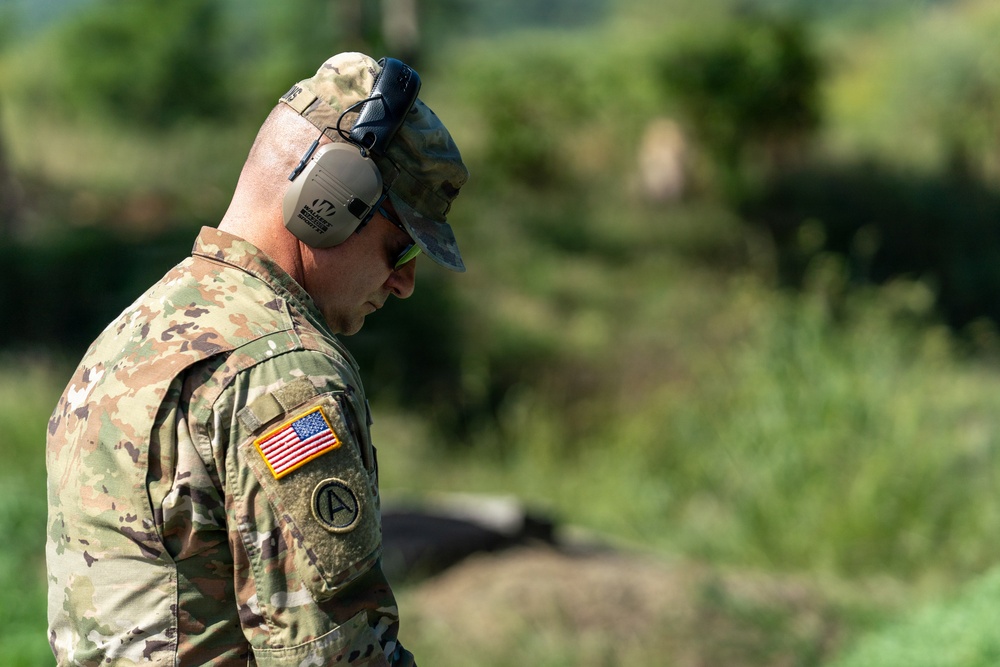 A U.S. Army soldier is photographed in profile view, looking downward, wearing full OCP (Operational Camouflage Pattern) uniform. The soldier is equipped with hearing protection earmuffs and safety glasses, suggesting participation in a live-fire range exercise. The background shows a lush, green outdoor environment consistent with a training range.