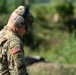 A U.S. Army soldier is photographed in profile view, looking downward, wearing full OCP (Operational Camouflage Pattern) uniform. The soldier is equipped with hearing protection earmuffs and safety glasses, suggesting participation in a live-fire range exercise. The background shows a lush, green outdoor environment consistent with a training range.
