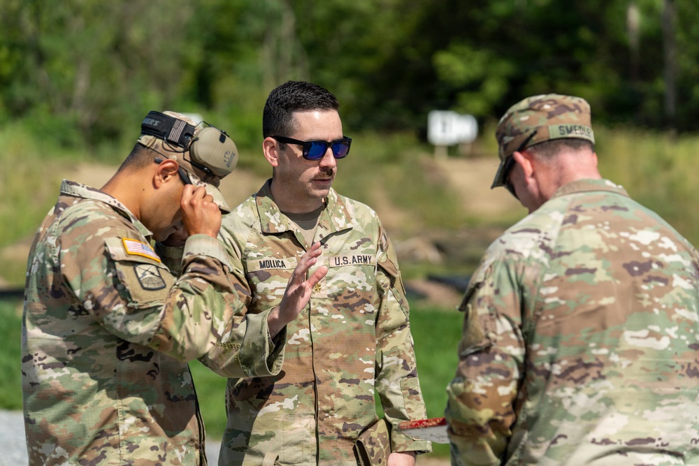 Three U.S. Army soldiers in OCP (Operational Camouflage Pattern) uniforms are engaged in a discussion at what appears to be a firing range or outdoor training area. The central figure, identified by name tape as 'MOLLICA,' appears to be briefing or instructing the others. The soldier on the left is wearing tactical hearing protection pushed up on his head.