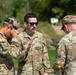 Three U.S. Army soldiers in OCP (Operational Camouflage Pattern) uniforms are engaged in a discussion at what appears to be a firing range or outdoor training area. The central figure, identified by name tape as 'MOLLICA,' appears to be briefing or instructing the others. The soldier on the left is wearing tactical hearing protection pushed up on his head.
