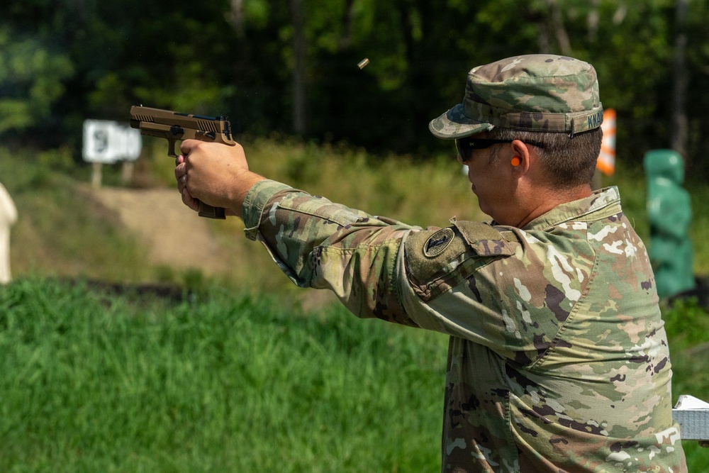 A U.S. Army soldier in Operational Camouflage Pattern (OCP) uniform is firing a pistol at an outdoor shooting range. A spent brass casing is visibly ejecting from the firearm mid-shot. The setting appears to be a military qualification or training range with target stands visible in the background.