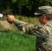 A U.S. Army soldier in Operational Camouflage Pattern (OCP) uniform is firing a pistol at an outdoor shooting range. A spent brass casing is visibly ejecting from the firearm mid-shot. The setting appears to be a military qualification or training range with target stands visible in the background.