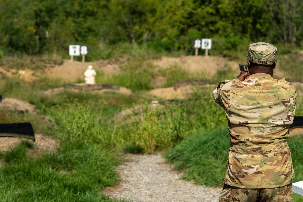 A soldier wearing OCP (Operational Camouflage Pattern) uniform with the name tape 'LATTA' is firing a pistol at a military shooting range. Multiple targets are visible downrange, including a 3D humanoid target in the foreground and numbered flat targets (78 and 89) in the background. The setting is an outdoor range with gravel pathways, earthen berms, and surrounding vegetation.