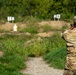 A soldier wearing OCP (Operational Camouflage Pattern) uniform with the name tape 'LATTA' is firing a pistol at a military shooting range. Multiple targets are visible downrange, including a 3D humanoid target in the foreground and numbered flat targets (78 and 89) in the background. The setting is an outdoor range with gravel pathways, earthen berms, and surrounding vegetation.