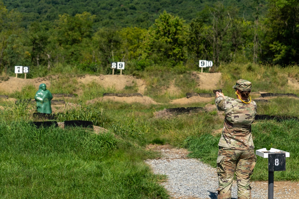 A soldier in full camouflage uniform is firing a pistol at a military shooting range, standing at lane marker number 8. The range features multiple numbered target positions (7-8, 8-9, 9-10) with a green human-silhouette target visible in the foreground. The setting is an outdoor range surrounded by dense green vegetation and wooded hills in the background.