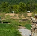 A soldier in full camouflage uniform is firing a pistol at a military shooting range, standing at lane marker number 8. The range features multiple numbered target positions (7-8, 8-9, 9-10) with a green human-silhouette target visible in the foreground. The setting is an outdoor range surrounded by dense green vegetation and wooded hills in the background.