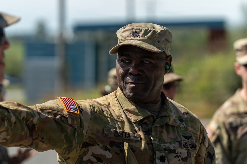 A U.S. Army officer in MultiCam/OCP camouflage uniform is photographed outdoors, appearing to gesture or point with his right arm extended. The soldier wears a patrol cap with what appears to be a Lieutenant Colonel oak leaf insignia, and a name tape reading 'DATTA' is visible on the uniform. The image is taken in a bright outdoor setting with other military personnel visible in the blurred background.