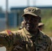 A U.S. Army officer in MultiCam/OCP camouflage uniform is photographed outdoors, appearing to gesture or point with his right arm extended. The soldier wears a patrol cap with what appears to be a Lieutenant Colonel oak leaf insignia, and a name tape reading 'DATTA' is visible on the uniform. The image is taken in a bright outdoor setting with other military personnel visible in the blurred background.