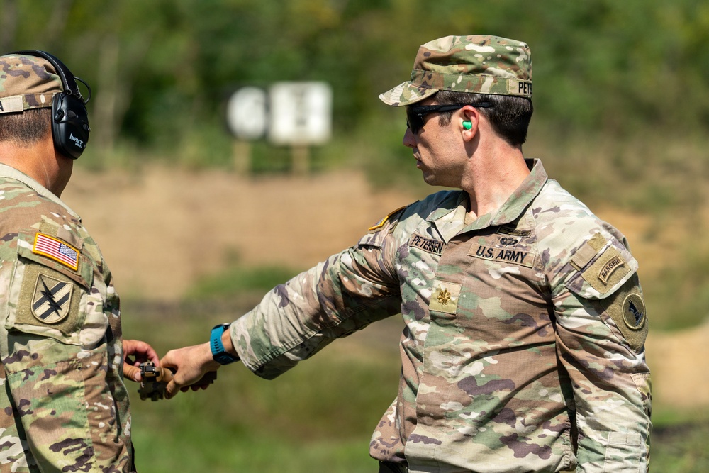 Two U.S. Army soldiers are at an outdoor firing range. The soldier facing the camera, identified by name tape as 'PETERSEN,' holds the rank of Major (gold oak leaf) and wears a Ranger tab. He appears to be handing or receiving a small object, likely a magazine or ammunition, from the other soldier.