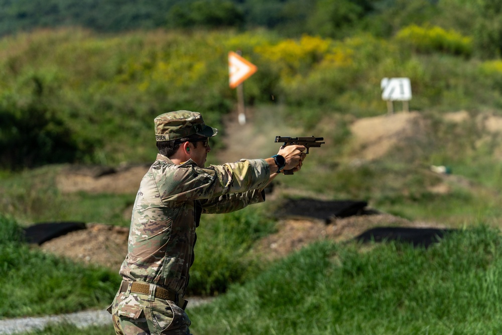 A soldier in U.S. Army OCP (Operational Camouflage Pattern) uniform is firing a pistol at an outdoor shooting range. The shooter is in a two-handed isosceles or modified Weaver stance with arms extended. Range markers and target indicators are visible in the background.
