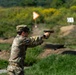 A soldier in U.S. Army OCP (Operational Camouflage Pattern) uniform is firing a pistol at an outdoor shooting range. The shooter is in a two-handed isosceles or modified Weaver stance with arms extended. Range markers and target indicators are visible in the background.