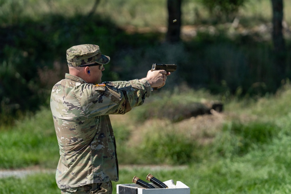A U.S. Army soldier in OCP (Operational Camouflage Pattern) uniform is firing a semi-automatic pistol at an outdoor shooting range. A spent brass casing is visibly ejecting from the firearm, indicating the weapon has just been discharged. Additional ammunition or magazines are visible on a white surface in the foreground.
