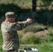A U.S. Army soldier in OCP (Operational Camouflage Pattern) uniform is firing a semi-automatic pistol at an outdoor shooting range. A spent brass casing is visibly ejecting from the firearm, indicating the weapon has just been discharged. Additional ammunition or magazines are visible on a white surface in the foreground.